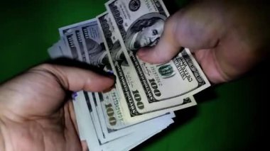 Loş bir odada, iki el hızlı bir para alışverişi yapıyor, 100 dolarlık banknotlar sergiliyor.. 
