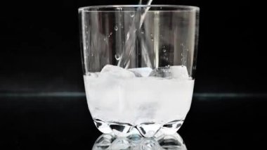 Karbonatlı içecek, siyah zemin üzerinde buz küpleri olan bir bardağa dökülüyor. Siyah yüzey üzerinde şeffaf bir içecekle doldurulmuş 4K 'lık bir içecek.