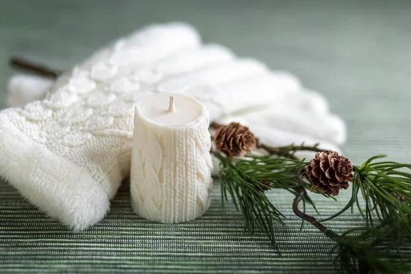A white candle with a knitted pattern, white knitted gloves and a pine branch with cones. Card. Photo