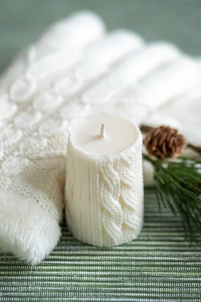 A white candle with a knitted pattern, white knitted gloves and a pine branch with cones. Card. Photo