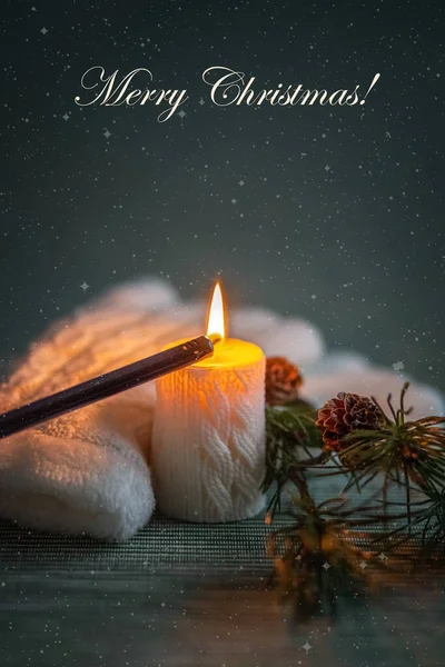 Lights the flame of a white candle with a knitted pattern and white knitted gloves and a pine branch with cones on a dark background. Snowfall. Card. Photo