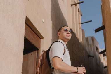 Sırt çantalı genç bir turist, güneş gözlüğü takıyor ve elinde bir fincan kahve ile Dubai 'deki eski kasabayı keşfediyor. Arap bir adamın portresi