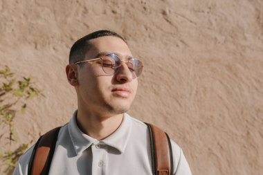 Güneş gözlüğü, tişört ve deri sırt çantası takan genç bir Arap 'ın portresi bej bir duvarın yanında gözleri kapalı ve sabah güneşinin tadını çıkarıyordu. Erkek öğrenci. Gen Z konsepti