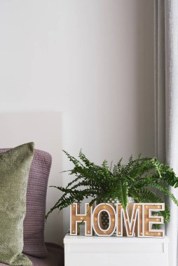 Yatak odasının içi: 'Ev' tabelası olan komodinli, yeşil ve mor yastıklı saksıda bulunan eğrelti otu ve yatak odası. Konforlu ev kavramı