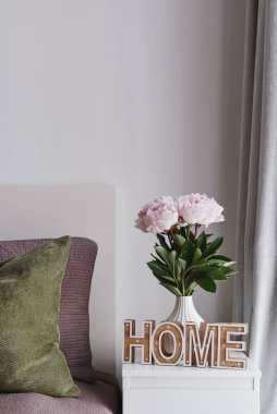 Konforlu ev kavramı. Pastel Pembe Şakayık Çiçekleri Beyaz Seramik Vazo ve Yatak Masasında Ev Tabelası