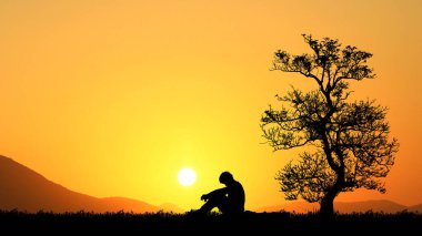 Yalnız adamın silueti turuncu gökyüzü arka planında, ağacın yanında oturuyor.