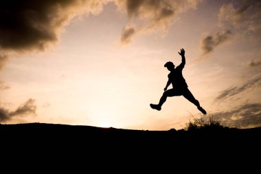 Gün batımında dramatik bir gökyüzüne doğru atlayan bir insanın silueti özgürlük ve macerayı simgeliyor..