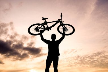 Çarpıcı bir siluet, dramatik bulutlarla dolu canlı bir günbatımı gökyüzüne karşı muzaffer bir şekilde bisiklet tutan bir kişi..