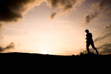 Gün batımında dramatik bir gökyüzü ve arka planda bulutlarla koşan bir insanın silueti açık hava fitness ve doğanın güzelliğini ön plana çıkarıyor..