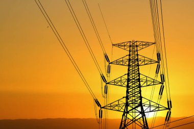 Yüksek voltajlı güç iletim kulesinin silüeti. Gün batımında enerji hatları olacak. Canlı turuncu gökyüzüne karşı kurulmuş..
