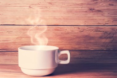 Beyaz bardakta dumanı tüten sıcak bir kahve, ahşap bir arka plana yaslanmış, sıcak bir sabah atmosferini çağrıştırıyor..