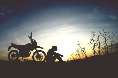 Gün batımında motosikletin yanında oturan sakin bir insan silueti. Arkasında çıplak ağaçlar ve renkli bir gökyüzü var..