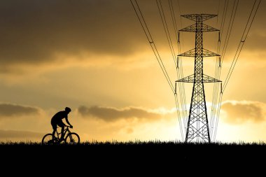 Gün batımında arka planda elektrik hatlarıyla bisiklet süren bir bisikletçinin silueti, altın saat boyunca açık hava macerasının ve enerji altyapısının özünü yakalıyor..