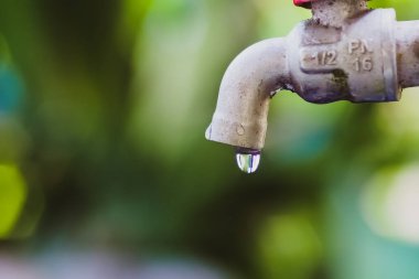 Bir damlası düşmek üzere olan bir musluğun ayrıntılı bir görüntüsü, canlı yeşil bulanık bir arkaplan üzerine kurulmuş..