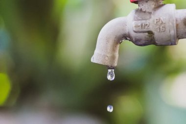 Su musluğunun ayrıntılı bir görüntüsü. Düşen bir damlacık, bulanık yeşil bir zemin üzerine kurulmuş. Suyun korunumunu vurguluyor..