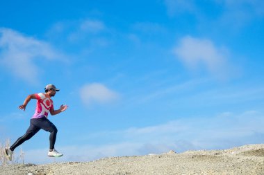 Spor giyimli atletik bir adam kayalık arazide açık mavi bir arka planda koşuyor, spor salonu ve açık hava aktivitesi sergiliyor..