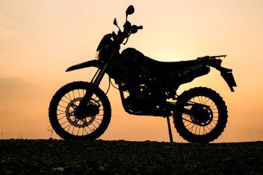 Motosikletin çarpıcı bir silueti altın bir günbatımı gökyüzüne yansıyor, çakıllı bir yolda motosikletin engebeli detaylarını vurguluyor..