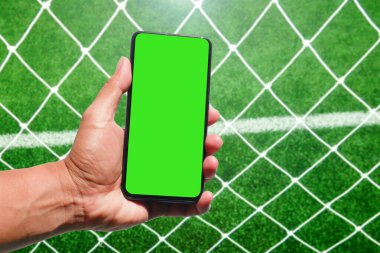 Futbol sahasının önünde yeşil ekranlı akıllı telefonu tutan ve ağ ören bir el. Spor ve teknoloji temaları için mükemmel..