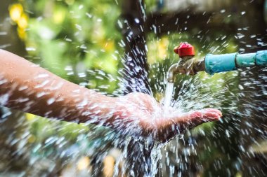 Açık bir musluktan bir ele su sıçrayan yakın plan, canlı, güneşli bir bahçe arka planına karşı kurulmuş..
