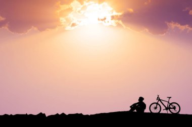 Canlı bir gün batımına karşı silueti olan, dramatik bulutlarla dolu bir bisikletin yanında oturan bir insanın sakin bir sahnesi..