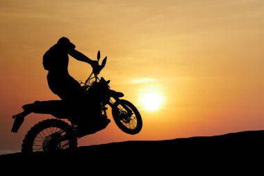 Motosikletlinin silueti arka planda canlı bir günbatımı, macera ve heyecan dolu bir manzarayla motosiklet sürüyor..