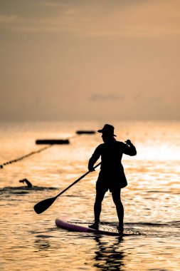 Gün batımında kürek çeken bir insanın sakin bir sahnesi, altın gökyüzüne bir siluet serpiştirip, okyanus sularını sakinleştiriyor..