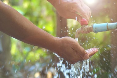 Arka planda su sıçratan, güneş ışığı ve yeşil yapraklı bir musluğun altında yıkanan ellerin yakın çekimi..