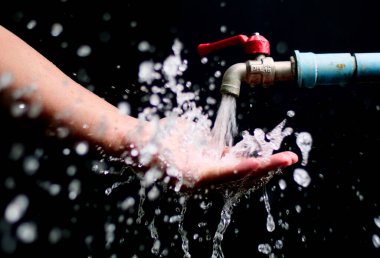 Musluktan akan suyun altında, su damlacıklarının siyah arkaplana sıçrayan bir elin yakın plan görüntüsü..