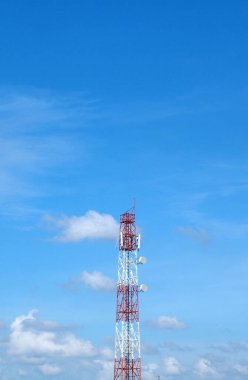 Uzun, kırmızı ve beyaz bir iletişim kulesi açık mavi bir gökyüzüne karşı duruyor. Güneşli bir günde dağınık beyaz bulutlar..