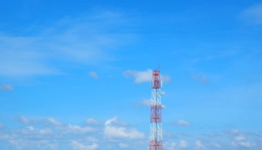 Uzun, kırmızı ve beyaz bir iletişim kulesi açık mavi bir gökyüzünün arkaplanında duruyor dağınık bulutlarla..
