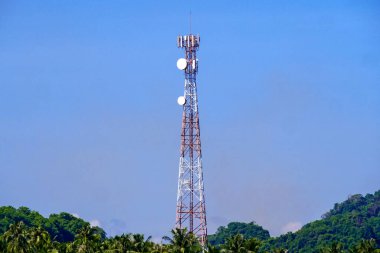 Önünde yemyeşil ağaçlar olan açık mavi gökyüzüne karşı duran yüksek bir iletişim kulesi..