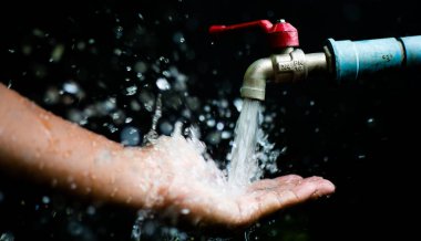 Akan bir musluğun altındaki bir elin yakın plan görüntüsü, su sıçrıyor ve damlacıklar koyu bir arkaplanda görünüyor..