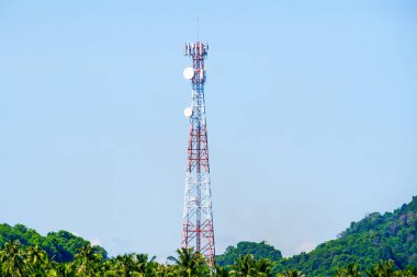 Açık mavi gökyüzü ve yemyeşil ağaçları olan tropikal bir arazinin ortasında duran uzun bir telekomünikasyon kulesi..