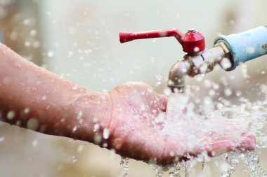 Açık hava musluğundan akan suyun altındaki bir elin yakın plan görüntüsü. Gün ışığında su damlacıklarını yakalıyor..