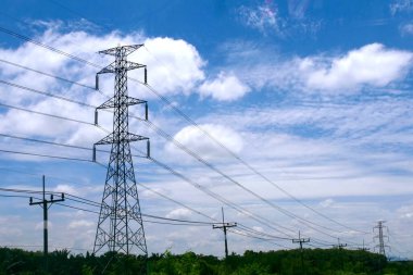 Enerji hatları parlak mavi bir gökyüzünün altında kabarık bulutlarla dolu yeşil bir arazi boyunca uzanıyor..