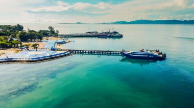 Berrak bir gökyüzü altında yemyeşil ve sakin mavi sularla çevrili, tekneli bir kıyı iskelesinin çarpıcı görüntüsü..