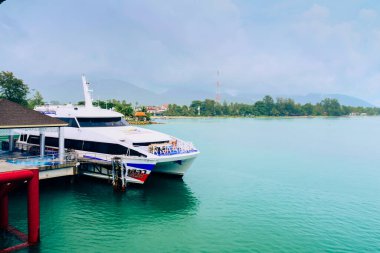 Modern bir feribot, turkuvaz suları ve berrak bir gökyüzünün altındaki yemyeşil sularla çevrili tropik bir ada iskelesine yanaşmış..