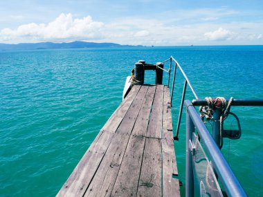 Sakin bir ahşap iskele, berrak mavi gökyüzünün altındaki uzak dağların çarpıcı manzarasını sunan canlı turkuaz okyanusa doğru uzanıyor..