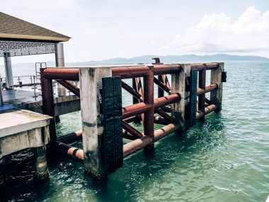 Paslı metal kirişli bir iskele okyanusa doğru uzanıyor, açık mavi bir gökyüzüne ve uzak dağlara karşı kuruluyor..