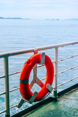 Canlı turuncu bir can simidi, arka planda sakin bir okyanus ve uzak adalar bulunan bir feribot korkuluğuna bağlı..