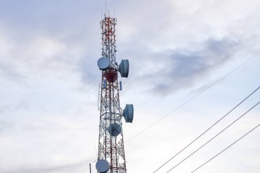 Uzun bir telekomünikasyon kulesi antenleri ve uydu antenleriyle bulutlu bir gökyüzüne kurulmuş, modern iletişim teknolojisi sergiliyor..