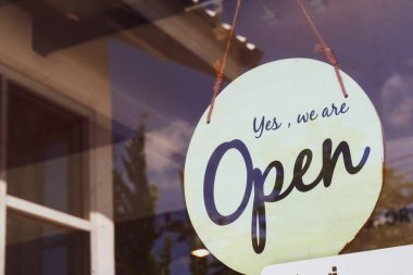 Bir dükkan kapısına asılmış klasik tarzda açık tabela, açık mavi gökyüzünü ve bulutları yansıtıyor..