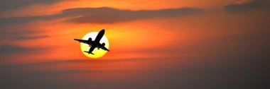 Canlı bir günbatımı gökyüzünde uçan bir uçak siluetinin çarpıcı görüntüsü dramatik renkleri ve bulut oluşumlarını vurguluyor.