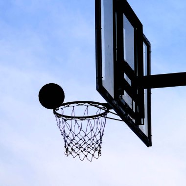 Mavi gökyüzüne karşı hareket eden bir topu olan basketbol potası silueti açık hava sporlarını ve eğlenceyi simgeliyor..