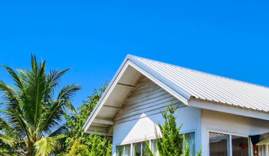 Parlak mavi gökyüzünün altında yemyeşil tropikal bitkilerle çevrili beyaz çatılı çağdaş bir ev..