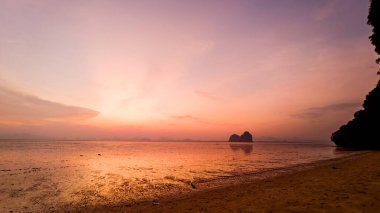 Sakin bir sahilde nefes kesici bir gün batımı, canlı renkler ve siluetli kaya oluşumları sakin sulara yansıyor..