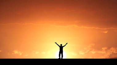 Kolları havada, çarpıcı turuncu bir gün batımına karşı dizilmiş, özgürlük ve neşe hissi uyandıran bir insan silueti..