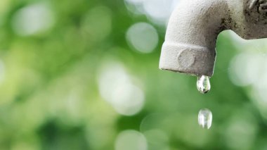 Su damlasının bir musluktan düşüşünün detaylı bir görüntüsü, yemyeşil bir bokeh arkaplan üzerine kurulmuş, korunmayı sembolize eden.