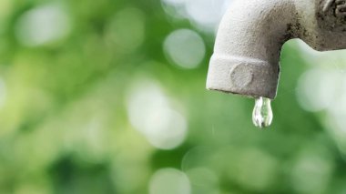 Bir musluktan düşen bir su damlasının detaylı bir görüntüsü, yemyeşil bir bokeh arka plan üzerine kurulmuş.