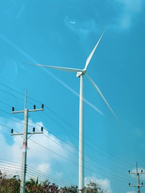 Rüzgâr türbini açık mavi gökyüzüne karşı dimdik durur. Önünde elektrik hatları olan yenilenebilir enerjiyi sembolize eder..